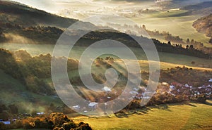 Aerial view of village in fog with golden sunbeams at sunrise in autumn. Beautiful rural landscape with road, buildings, foggy