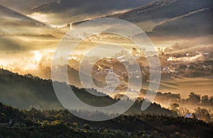 Aerial view of village in fog with golden sunbeams at sunrise in autumn. Beautiful rural landscape with road, buildings, foggy