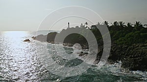 Aerial view of waves crashing on rocks near lighthouse, beacon for ships at sea, surrounded by lush foliage on hill