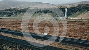 Aerial view of a white car driving along a road with a picturesque waterfall in the background