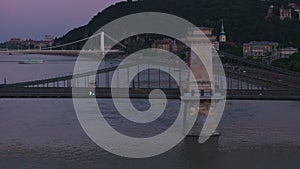 Aerial view of wide Danube river and its bridges at dusk. Cars driving on waterfront road. Budapest, Hungary