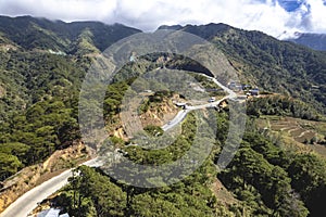 Aerial view of a winding road cutting through a lush mountain landscape with scattered settlements and vehicles. At the boundary