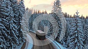 Aerial view of winter landscape with snow covered mountain hills and winding forest road with driving vehicles in morning.