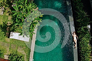 Aerial view of woman in beige bikini relaxing and sunbathe on edge of infinity pool around palm trees and jungle. Luxury