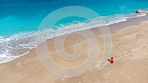 Aerial view of woman in red dress sitting on the sandy beach with a baby, enjoying soft turquoise ocean wave. Tropical sea in