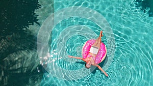 Aerial view of a young brunette woman swimming on an inflatable big donut with a laptop in a transparent turquoise pool.