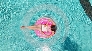 Aerial view of a young brunette woman swimming on an inflatable big donut with a laptop in a transparent turquoise pool.