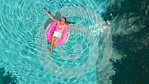 Aerial view of a young brunette woman swimming on an inflatable big donut with a laptop in a transparent turquoise pool.