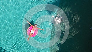 Aerial view of a young brunette woman swimming on an inflatable big donut with a laptop in a transparent turquoise pool.