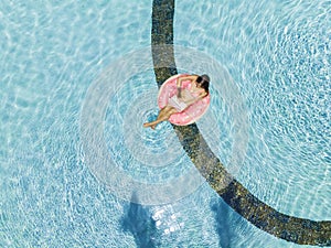 Aerial view of a young brunette woman swimming on an inflatable big donut with a laptop in a transparent turquoise pool.