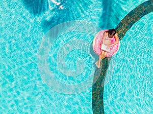 Aerial view of a young brunette woman swimming on an inflatable big donut with a laptop in a transparent turquoise pool.