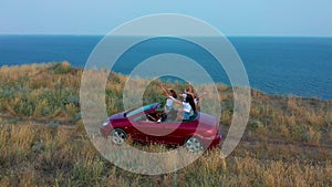 Aerial view. Young girls driving convertible car at sunset by the sea front beach. Summer adventure.