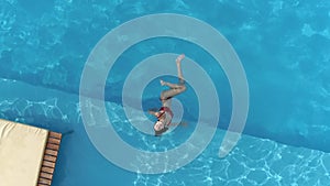 Aerial view, young happy woman relaxes in blue swimming pool at sunny day and looks at camera