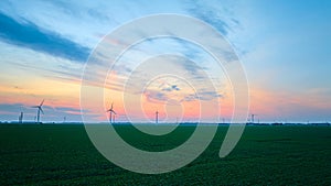 Aerial wide shot of wind farm beyond green farmland on gorgeous sunset evening