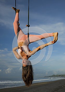 Aerial yoga beach workout - young attractive and healthy woman practicing aero-yoga training balance body and mind control hanging