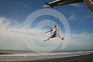 Aerial yoga beach workout - young attractive and healthy woman practicing aero-yoga training balance body and mind control hanging