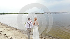 Aero, beautiful newlyweds walking along the beach, under a transparent umbrella, against the blue sky, river, and a