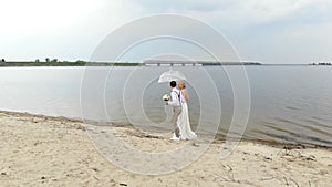 Aero, beautiful newlyweds walking along the beach, under a transparent umbrella, against the blue sky, river, and a