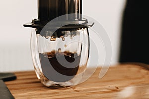 Aeropress and glass cup, spoon on wooden table, coffee drops closeup in glass. Professional barista preparing coffee alternative