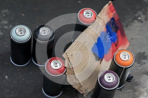 Aerosol cans of spray paint and cardboard palette for paint, graffiti on a dark background of asphalt. Top view. Urban