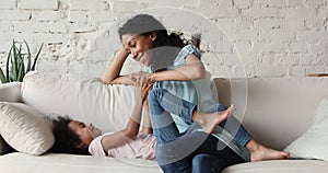 Affectionate Black mom messing on sofa with playful little daughter