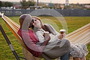 Affectionate couple embraces while sitting in hammock outdoors, wrapped in warm blanket. Black man and Caucasian woman deeply in