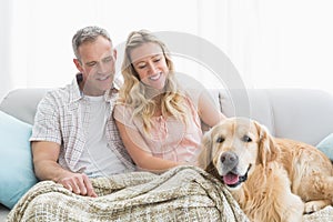 Affectionate couple sitting on sofa under blanket with their dog