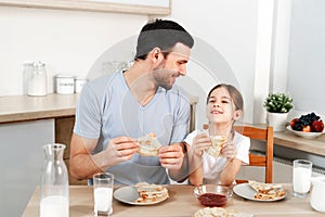 Affectionate pleasant looking father and his little daughter eat tasty morning meal at kitchen, communicate with each