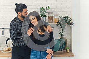 Affectionate scene between two people in the kitchen. Proud young bearded handsome Cuban man hugging his long-haired