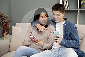 Affectionate young couple with mobile phones relaxing on sofa. Happy diverse woman and man hugging using smartphone