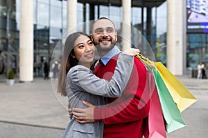 Affectionate young international couple with shopper bags smiling and hugging near big mall