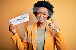 African American afro businesswoman with curly hair holding paper with capitalism message happy with big smile doing ok sign,