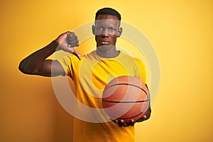 African american athlete man holding basketball ball standing over  yellow background with angry face, negative sign