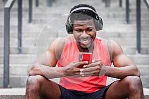 African american athletic guy listens to music in wireless headphones and smiles outdoors, a man sits on the steps and looks into