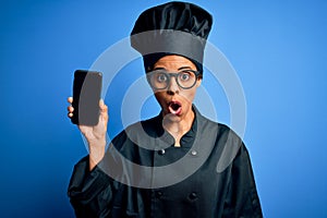 African american baker woman wearing cooker uniform holding smartphone showing screen scared in shock with a surprise face, afraid