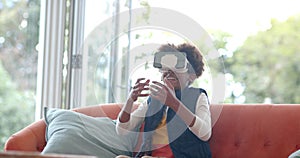 African american boy sitting on sofa, using vr headset and touching virtual screen, slow motion