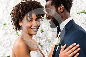 African american bride hugging handsome bridegroom near flowers