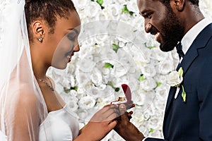 African american bride looking at box with ring near happy bridegroom and flowers