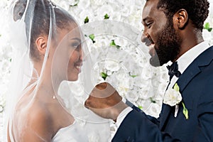 African american bridegroom touching white veil and smiling near bride and flowers