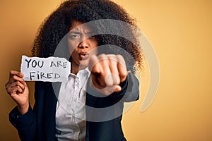 African american business boss woman with afro hair holding you are fired paper for dismissal pointing with finger to the camera