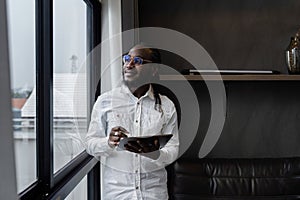 African American business man working at home on weekend, holding tablet and standing looking out the window