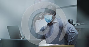 African american businessman in face mask talking on smartphone, using laptop in conference room