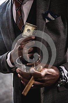 African american businessman holding glass of alcohol beverage and cigar while hiding dollar banknote