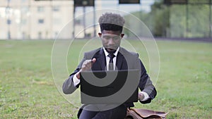 African american businessman sitting near office outside, giving thumb up, while using laptop