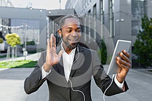 African american businessman in a suit and headphones standing outside an office center and talking on a video call from