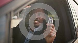 African American businessman in suit sitting inside his luxury electric car with keys in hands. Handsome man looks