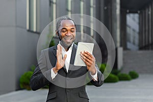 An African American businessman in a suit stands outside an office center and talks on a video call. He holds a tablet