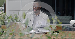 African American businessman working with documents and using laptop outside office center in street cafe