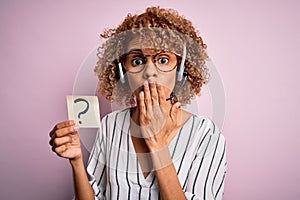 African american call center agent woman using headset holding paper with question mark cover mouth with hand shocked with shame