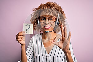 African american call center agent woman using headset holding paper with question mark doing ok sign with fingers, excellent